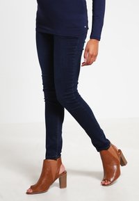 Tmavě modré skinny džíny se hladkým povrchem, doplněné o béžové open-toe kotníkové boty na podpatku s blokovým podpatkem a výřezovým designem.