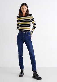Striped long-sleeve top in black, olive, and yellow; paired with dark blue jeans and black lace-up boots. Smooth fabric with fitted design.