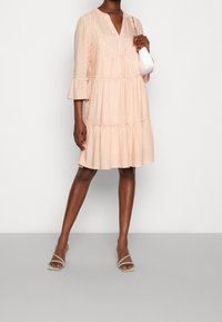 Vestido de rayas rosa claro con escote en V, mangas acampanadas de tres cuartos y dobladillo en capas. Combinado con sandalias verdes con tiras y un pequeño bolso blanco.