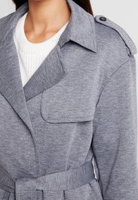 Šedý trenčkot z měkké látky s výrazným límcem, zapínáním na ramenních akcentech a páskem v pase. Na přední straně je našitá kapsa.