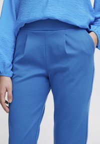 Pantalons bleus avec une texture lisse, présentant des plis à l'avant, une taille haute et des poches latérales. Le tissu a une apparence douce et unie.
