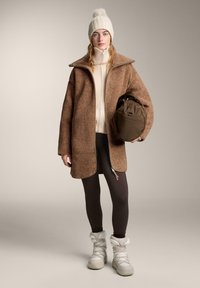 Barna gyapjúkabát magas gallérral, alatta krémszínű pulóver, sötét leggings, és fehér szigetelt csizma; sötétbarna táskát fog a kezében.