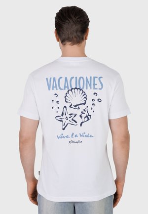 Man draagt een wit T-shirt met blauwe schelpen en de tekst "VACACIONES" en "Viva La Vida" op de achterkant, tegen een effen achtergrond.