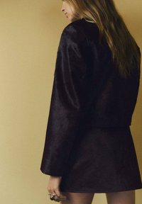 Giacca in pelliccia sintetica bordeaux scuro con un design corto e maniche ampie, abbinata a una mini gonna coordinata, su uno sfondo giallo tenue.