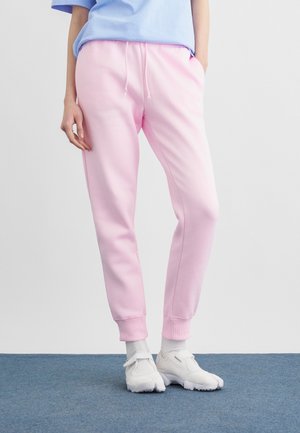 Personne portant un pantalon de jogging rose clair, des chaussettes blanches et des baskets blanches, debout sur un tapis bleu contre un mur blanc.