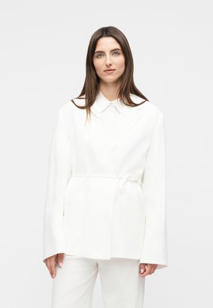 Kvinde med glat brunt hår iført en oversized hvid blazer med bindebælte og matchende hvide bukser, der kigger ind i kameraet.