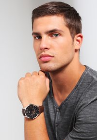 Montre à bracelet en cuir brun avec un cadran noir, des accents argentés et des index horaires visibles, posée sur une chemise grise texturée.