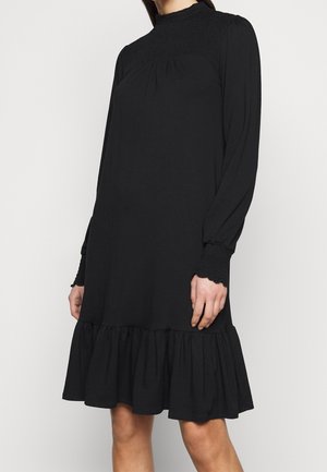 Vestito nero a maniche lunghe fino al ginocchio con collo alto arricciato, polsini arricciati e orlo con volant indossato da una persona in piedi.