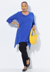 Blaues Tunika-Oberteil mit V-Ausschnitt und Dreiviertelärmeln, kombiniert mit schwarzen Leggings. Hält eine strukturierte gelbe Handtasche und trägt schwarze Ballerinas.