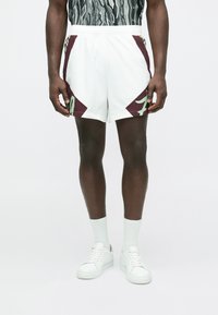 Balti sporta šorti ar bordo un zaļu ģeometrisku akcentu, elastīga josta, valkāti kopā ar baltām zeķēm un sporta apaviem. Gluda auduma tekstūra.