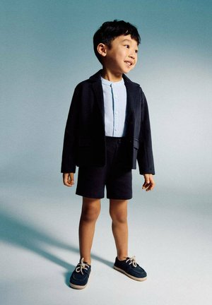 Mlad fant se smeje in stoji, oblečen v mornarsko modri blazer, svetlo modro srajco z gumbi, mornarsko modre hlače in mornarsko modre čevlje z belimi vezalkami.