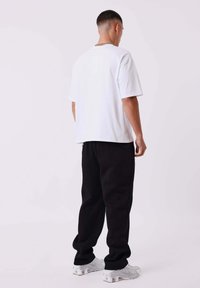 Uomo di spalle che indossa una t-shirt bianca, pantaloni neri, sneakers bianche e una collana in argento su uno sfondo neutro.