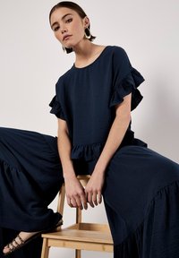 Navy blue textured top with ruffle sleeves and matching wide-leg pants. Wooden chair; model has short hair and minimal accessories.