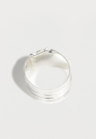 Ευρύ ασημένιο δαχτυλίδι με υφασμένα οριζόντια μπάντα και άνισες, οργανικές άκρες που ακουμπούν σε μια απλή λευκή επιφάνεια.