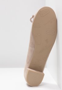 Ballerine beige en daim avec bout arrondi, nœud décoratif et semelle plate en caoutchouc ornée de motifs et de marquages en relief.