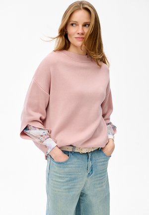 Młoda kobieta o jasnobrązowych włosach, ubrana w luźny różowy sweter z kwiatowymi rękawami, jasnoniebieskie dżinsy i beżowy pasek, z rękami w kieszeniach.