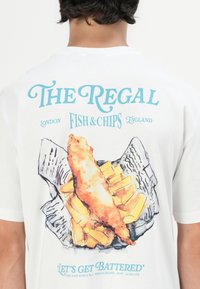 White t-shirt featuring colorful graphic design of fish and chips, with text "The Regal Fish & Chips England" and "Let's Get Battered" below.