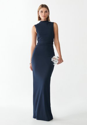 Vrouw in een mouwloze marineblauwe, vloer-lange jurk die een kleine zilveren clutch vasthoudt, staand tegen een effen witte achtergrond.