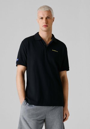 Giovane uomo con capelli biondi corti che indossa una polo nera "Red Bull Racing" e pantaloni grigi, in piedi con una mano in tasca.