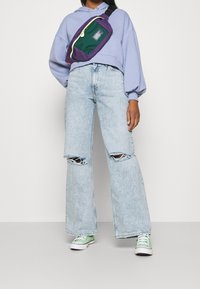 Violetti ja vihreä olkalaukku vetoketjulla, joka on pukeutunut vaaleanviolettiin huppariin ja kuluneisiin vaaleansinisiin leveälahkeisiin farkkuihin, yhdistettynä vihreisiin lenkkareihin.