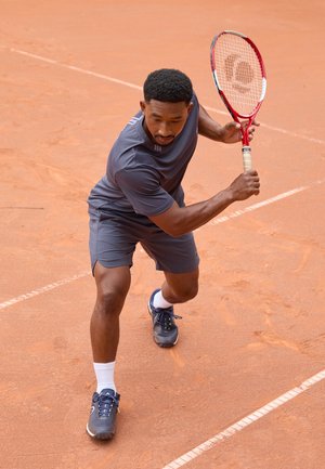Tennis spiller iført gråt træningstøj, holder en rød racket, på en grusbane. Bemærkelsesværdige detaljer: korte ærmer, sportssko og hvide sokker.
