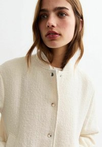Crème kleurige blazer met een hoge kraag, voorzien van zilveren drukknoopjes en zijzakken. De stof heeft een zachte, geweven uitstraling.