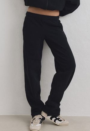 Persona con pantalones deportivos negros y zapatillas blancas con rayas negras, de pie contra una pared clara, recortada a la altura de la cintura.