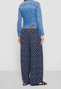 Persona vistiendo una chaqueta de mezclilla azul con pantalones anchos de estampado azul marino y sandalias negras, de pie frente a un fondo gris claro liso.