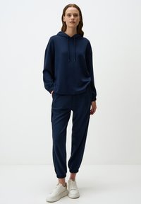 Haut à capuche bleu marine avec cordon de serrage ajustable et coupe ample, associé à un jogging assorti avec des poignets élastiques. Des baskets blanches complètent la tenue.