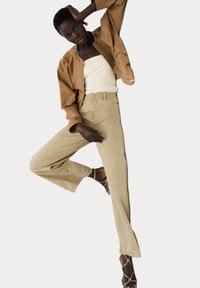 Hnědá bombera bunda přes bílý žebrovaný top, doplněná světle béžovými vykrojenými džíny a sandály na páscích. Hladké textury a čisté linie.