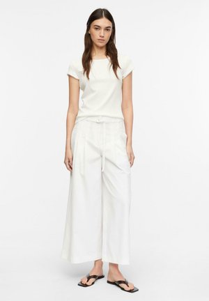 Kobieta ubrana w biały krótki top, białe szerokie spodnie z paskiem oraz czarne sandały typu japonki, stojąca na jednolitym tle.