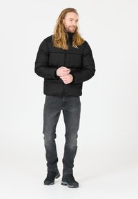Svart puffjacka med dragkedja, quiltad design och hög krage. Kombinerad med mörka jeans och svarta vandringsskor mot en neutral bakgrund.