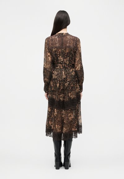 Γυναίκα με μακριά σκούρα μαλλιά φοράει ένα ημιδιάφανο, εμπριμέ φόρεμα μέχρι το μέσο της γάμπας με δαντελένιες λεπτομέρειες και μαύρες μπότες ψηλά στο γόνατο με τακούνια, στέκεται γυρισμένη με την πλάτη.