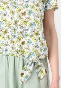 Bluse med blomsterdesign på hvit bakgrunn som har grønne, blå og brune blomster. Bundet i midjen, kombinert med lysegrønne bukser.