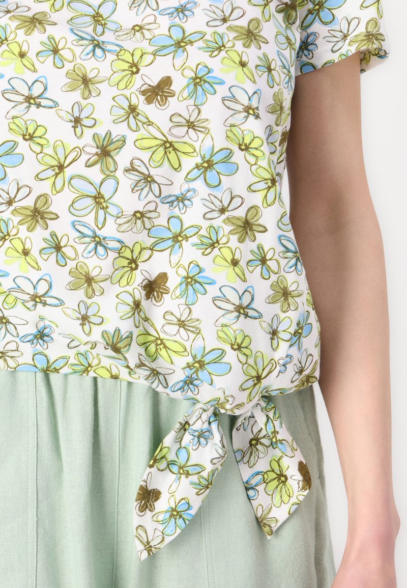 Blusa con motivo floreale su sfondo bianco, con fiori verdi, blu e marroni. Legata in vita, abbinata a pantaloni verde chiaro.