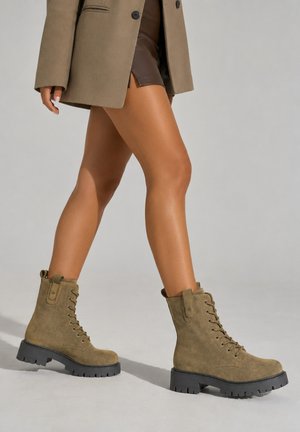 Gambe di una persona che indossa stivali da combattimento in camoscio marrone con lacci, una gonna corta marrone e un cappotto beige che cammina su uno sfondo uniforme.