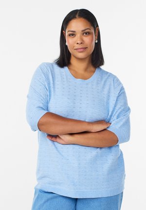 Giovane donna con carnagione media e capelli lisci neri, che indossa un maglione azzurro chiaro e jeans, con le braccia incrociate, guarda la fotocamera con sicurezza.