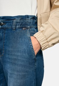 Denim jeans in medium blue with a relaxed fit. Features include a high waistband, side pockets, and a small brand label near the waist.