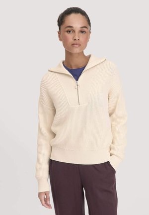 Crème-kleurige gebreide pullover met een kwart-rits, geribbelde textuur, brede mouwen en een aansluitende tailleband. Draagover een blauwe shirt.