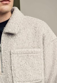 Šviesiai pilka flisinė zip-up striukė. Pasižymi tekstūruota paviršiumi, apykakle ir krūtine kišenę su siuvinėtais akcentais.