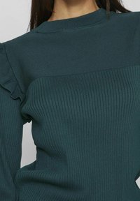 Femme portant un pull en tricot côtelé bleu sarcelle foncé avec un col rond haut et un petit détail volanté sur la couture de l'épaule.