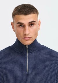 Pull-over marin en maille texturée avec col montant et détail de fermeture éclair en métal. Le tissu semble épais et chaud.