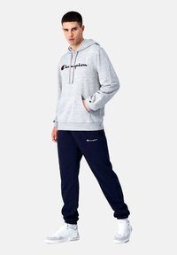 Grijze hoodie met een zwart scriptlogo, gecombineerd met marineblauwe sweatpants met een logo, en witte sneakers met grijze accenten.