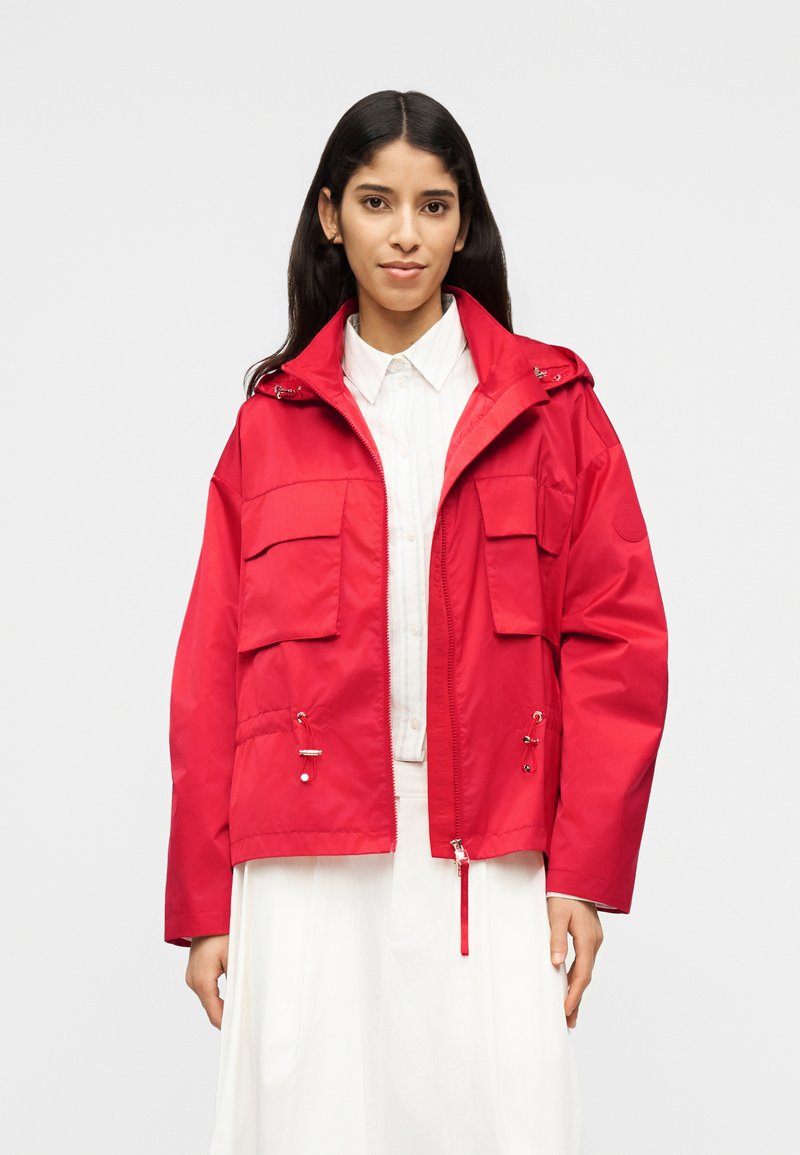 Γυναίκα με μακριά σκούρα μαλλιά, φορώντας κόκκινο μπουφάν με φερμουάρ και τσέπες με καπάκι πάνω από λευκό πουκάμισο με γιακά και φούστα, στέκεται μπροστά σε απλό φόντο.