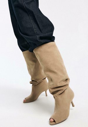 Beige suede slouchy knee-high boots with peep toes and slim heels worn with dark denim jeans.