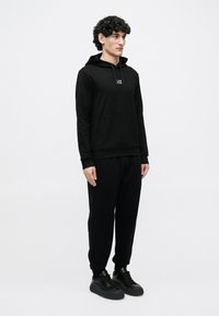 Joven de pie vistiendo una sudadera con capucha negra con logo, pantalones jogger negros y zapatillas negras sobre un fondo claro liso.