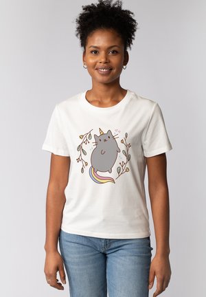 Femme souriante aux cheveux bouclés portant un t-shirt blanc avec un chat licorne gris, une queue colorée et des branches fleuries autour.