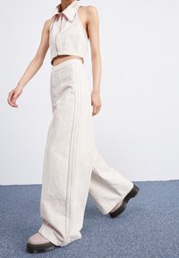 Kvinna i ljusbeige vida byxor och matchande kort ärmlös jacka, går på blåmattad golv, iklädd kraftiga svarta stövlar.