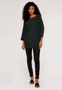 Oversized top i sort og grøn med pletter, bred halsudskæring og trekvartlange ærmer, parret med sort tætsiddende bukser og åbne sorte sko.
