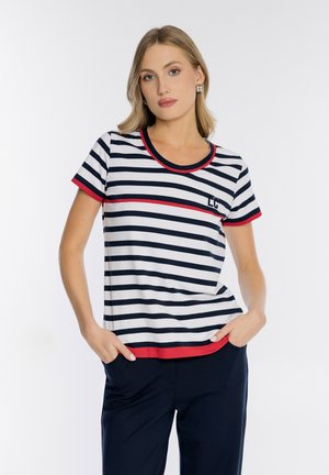 Kobieta w granatowo-białej koszulce w paski z czerwonym wykończeniem, stojąca z rękami w kieszeniach granatowych spodni, o neutralnym wyrazie twarzy, na jasnym tle.